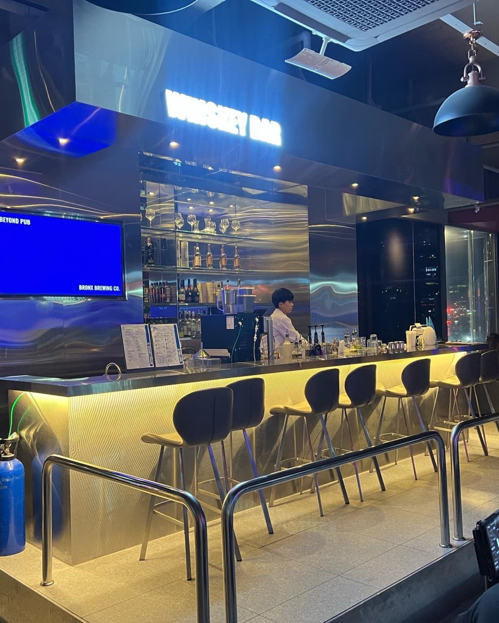 푸른 네온 아래 스테인리스 소재의 긴 카운터와 하단 조명이 켜진 1인용 바 좌석 전경