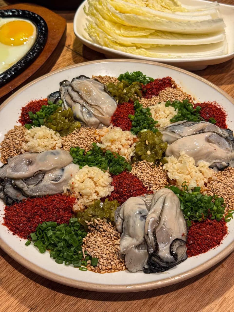 나무 테이블 위 고춧가루, 쪽파, 마늘 양념을 얹은 생굴 접시와 알배추, 계란후라이가 놓인 1인 안주 상차림