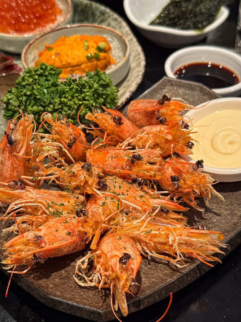 어두운 테이블 위 새우 머리 튀김과 성게알, 연어알이 담긴 작은 접시들이 놓인 1인용 안주 차림