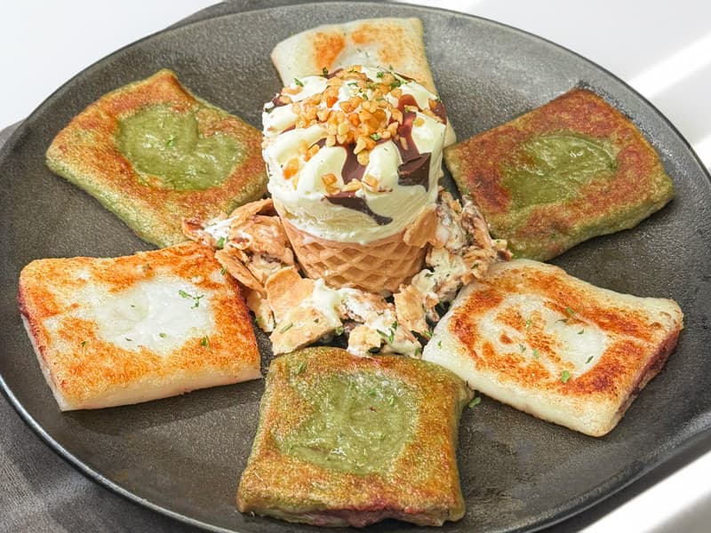 검은색 원형 접시 위에 놓인 녹색과 흰색의 구운 사각 떡과 초코 시럽을 곁들인 아이스크림 콘