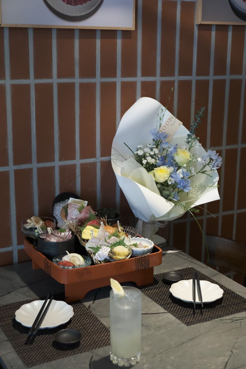 붉은 타일 벽 앞 테이블 위 사시미가 담긴 목재 트레이와 꽃다발, 레몬을 띄운 하이볼 잔과 1인용으로 배치된 식기 세트