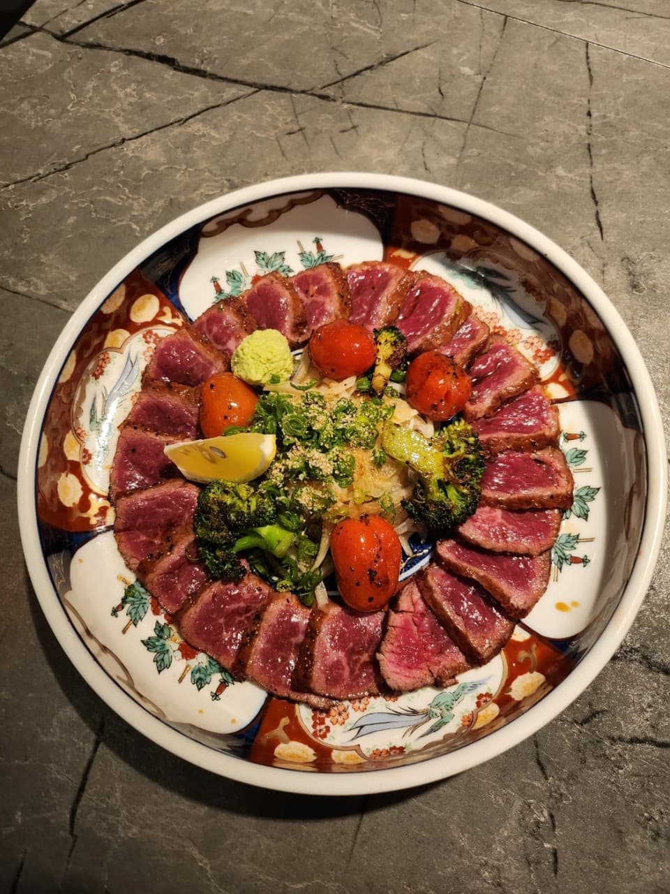 어두운 석재 테이블 위 문양 접시에 1인용으로 정갈하게 담긴 소고기 타다끼와 와사비, 구운 채소