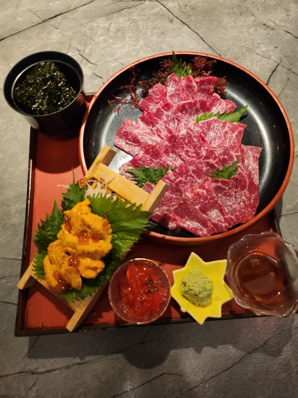 석재 질감 테이블 위 붉은 쟁반에 담긴 육사시미와 성게알, 김, 와사비로 구성된 1인용 술상 차림