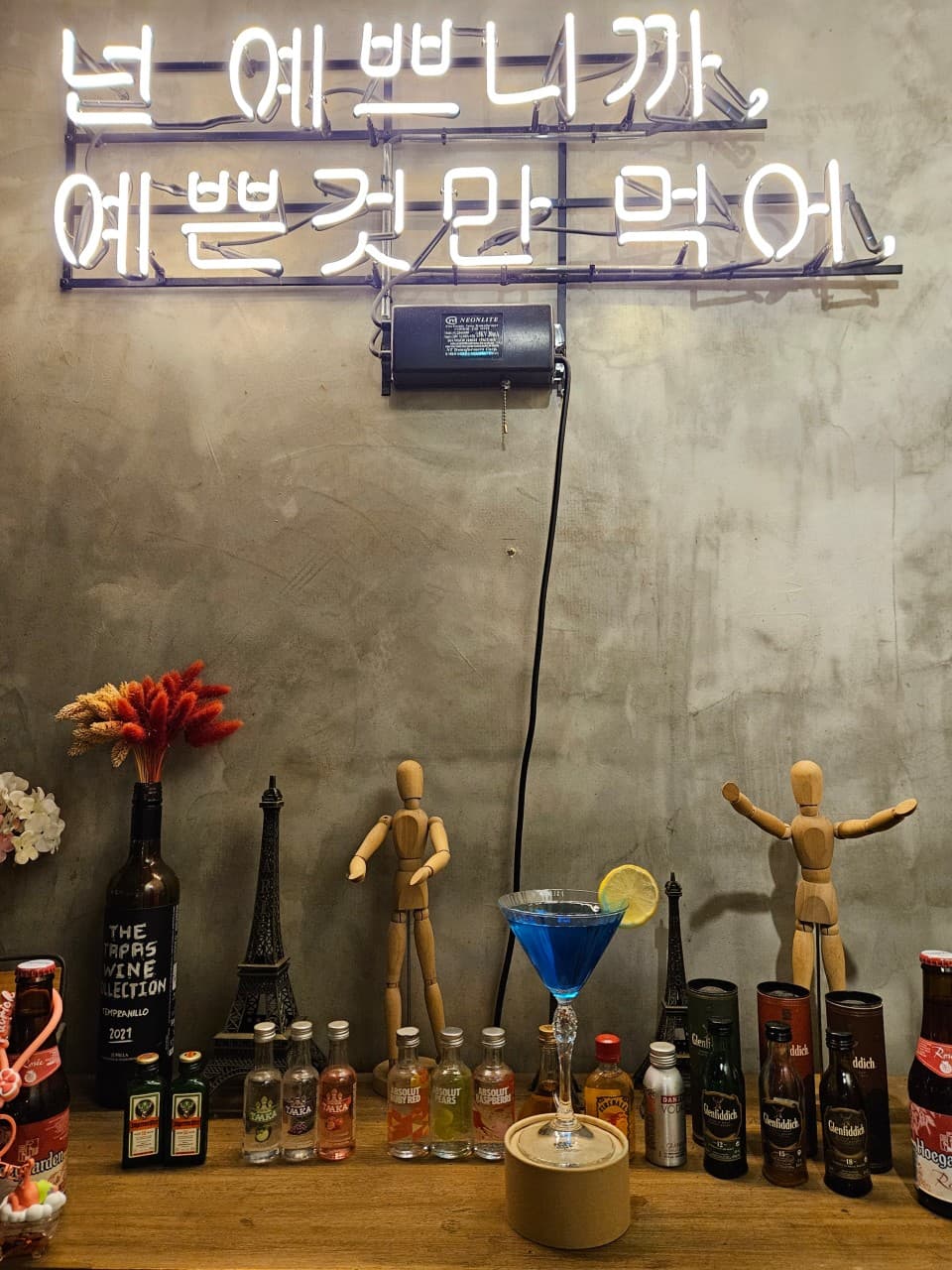 네온사인이 부착된 노출 콘크리트 벽면 아래 목재 카운터 위 푸른 칵테일과 미니어처 술병 및 소품들