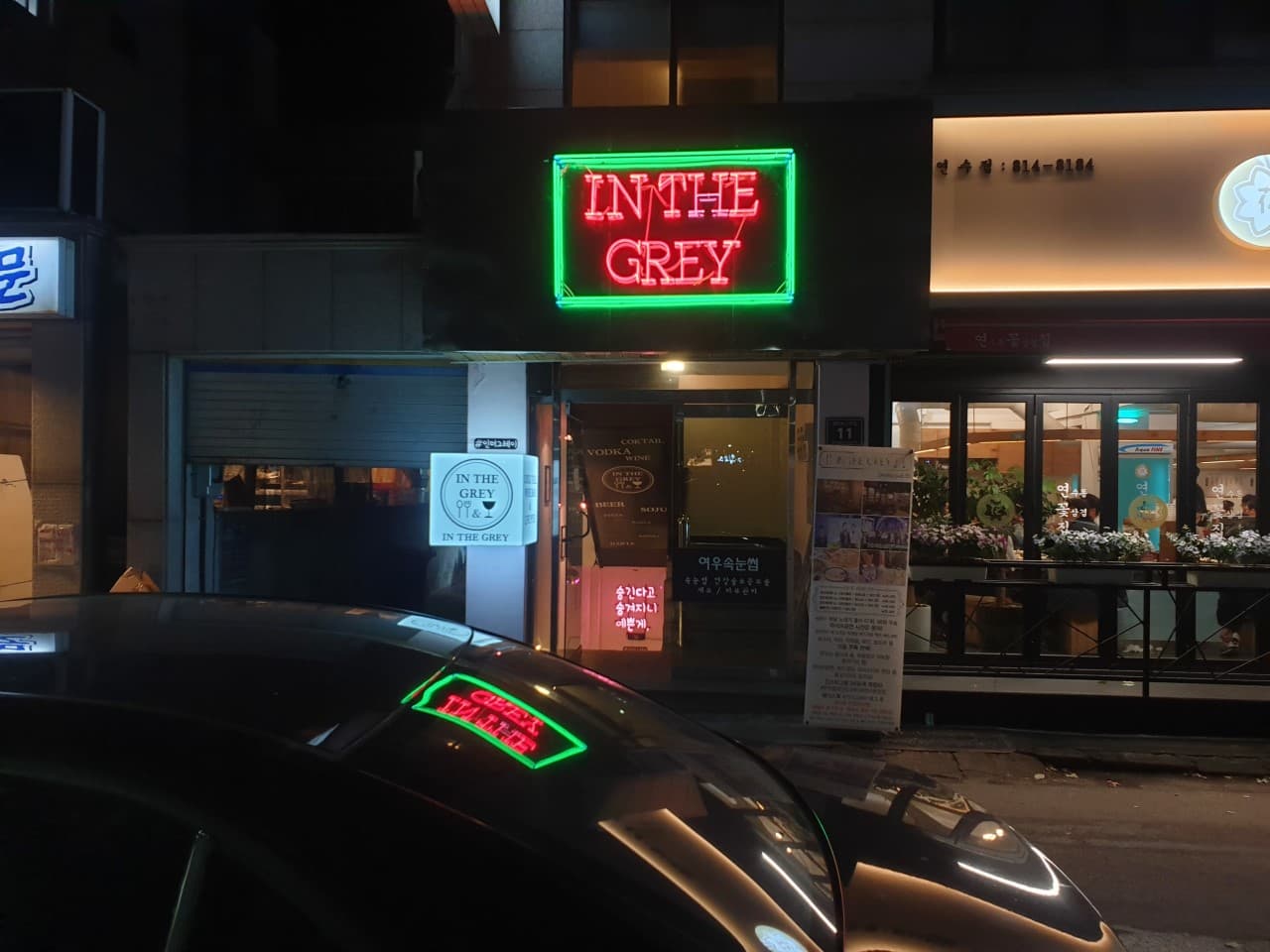 어두운 밤 붉은색과 초록색 네온사인이 켜진 건물 입구와 입간판. 자동차 보닛에 네온 조명이 반사되는 야간의 바 출입구 전경