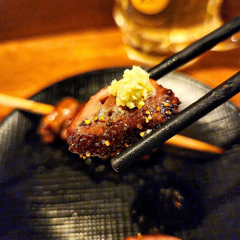 어두운 조명 아래 검은 젓가락으로 집어 든 와사비 얹은 닭꼬치 한 점과 배경의 하이볼 잔