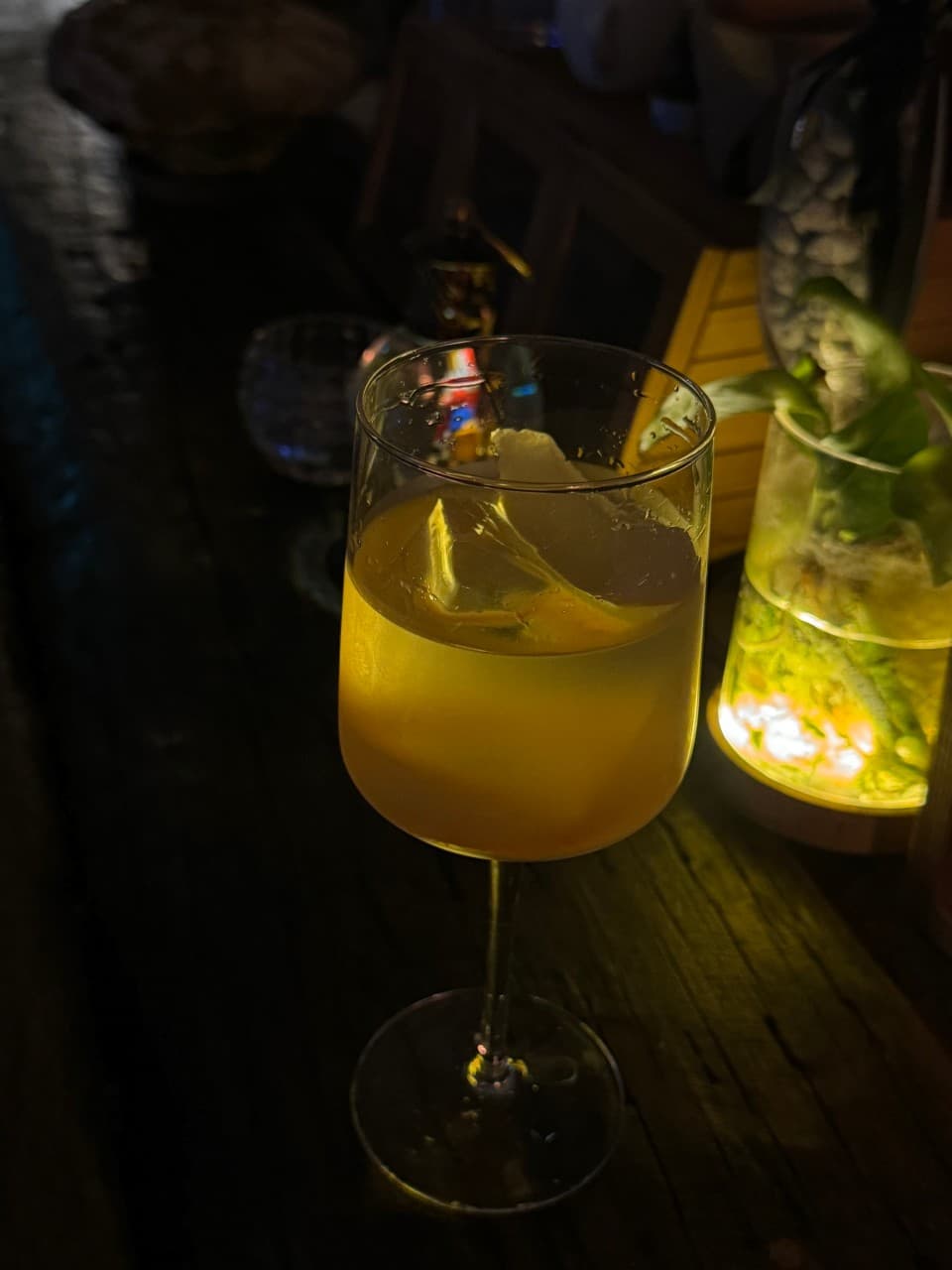 어두운 조명 아래 목재 바 테이블 위 얼음 가득한 노란색 술잔과 조명을 받는 작은 식물 화분