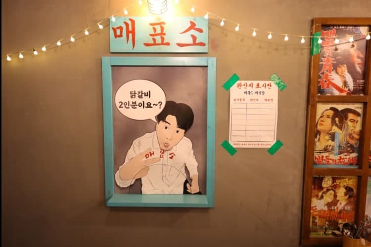 매표소 간판 아래 캐릭터 일러스트와 고전 영화 포스터가 부착된 어두운 톤의 벽면과 주황빛 줄전구가 빛나는 공간