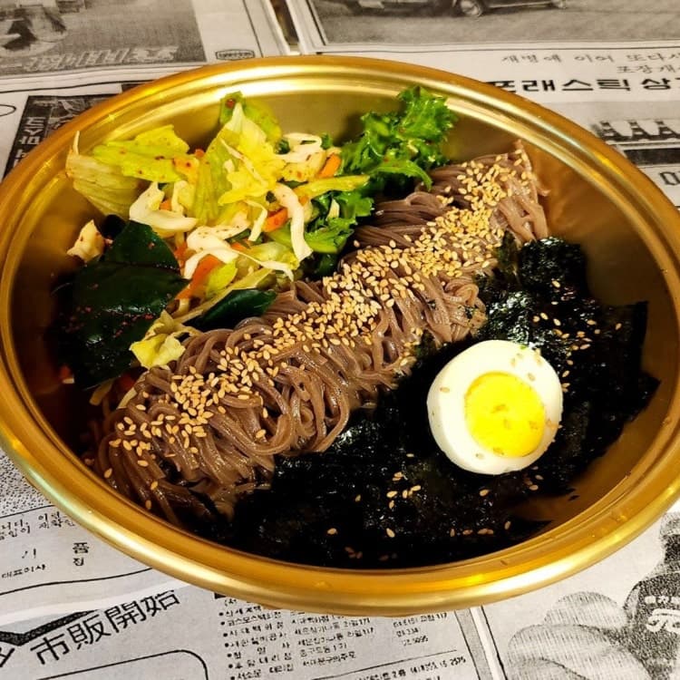 신문지 패턴의 매트 위 금색 그릇에 담긴 메밀면과 녹색 채소, 김가루, 삶은 계란 반 알