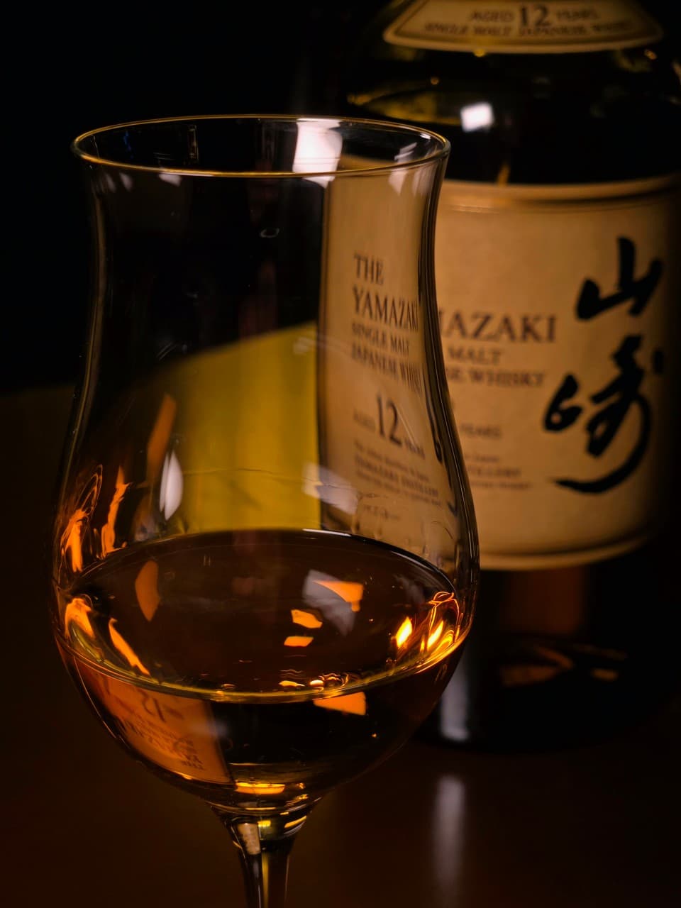 어두운 조명 아래 위스키가 담긴 튤립형 잔과 그 뒤에 배치된 야마자키 12년 위스키 병