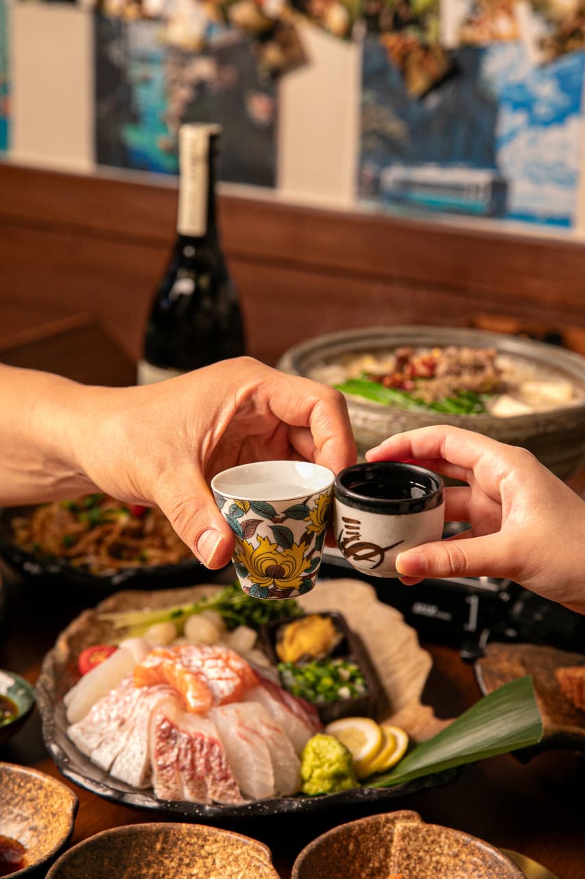 작은 도자기 술잔을 맞댄 두 손 아래 모둠 사시미와 전골 냄비, 술병이 놓인 어두운 나무 질감의 테이블