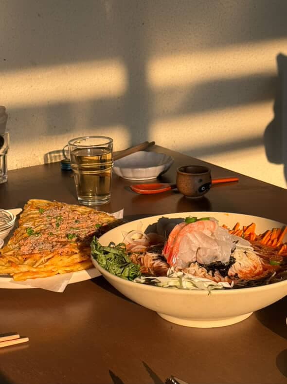 석양 빛이 비치는 테이블 위 새우와 회를 얹은 비빔면, 노란 감자채전과 술이 담긴 투명한 유리잔