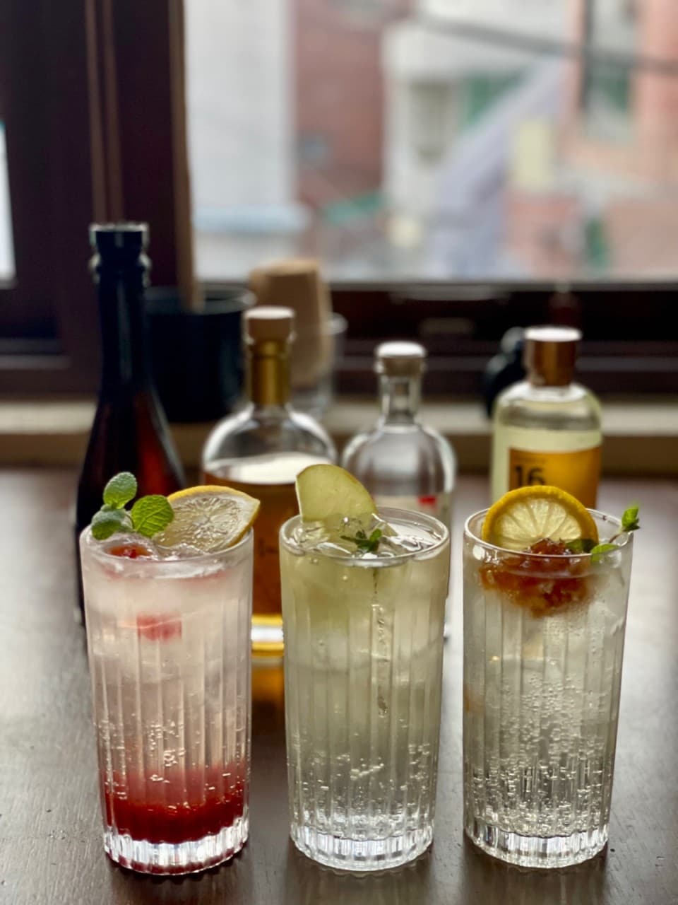 창가 나무 다찌석 위 레몬과 민트, 사과 조각이 담긴 세 잔의 하이볼과 배경에 나열된 다양한 술병