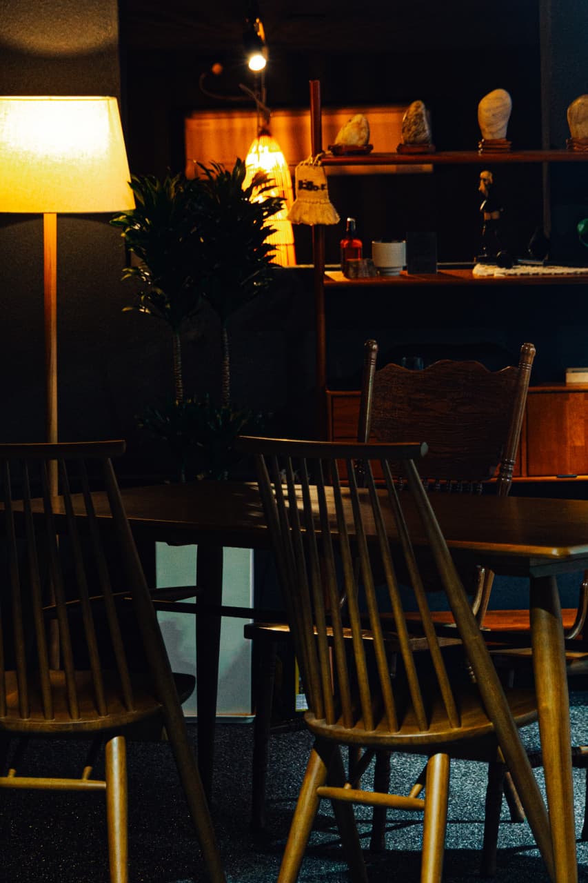 어두운 실내의 노란 스탠드 조명 아래 배치된 원목 테이블과 1인용 등받이 의자 및 장식 선반