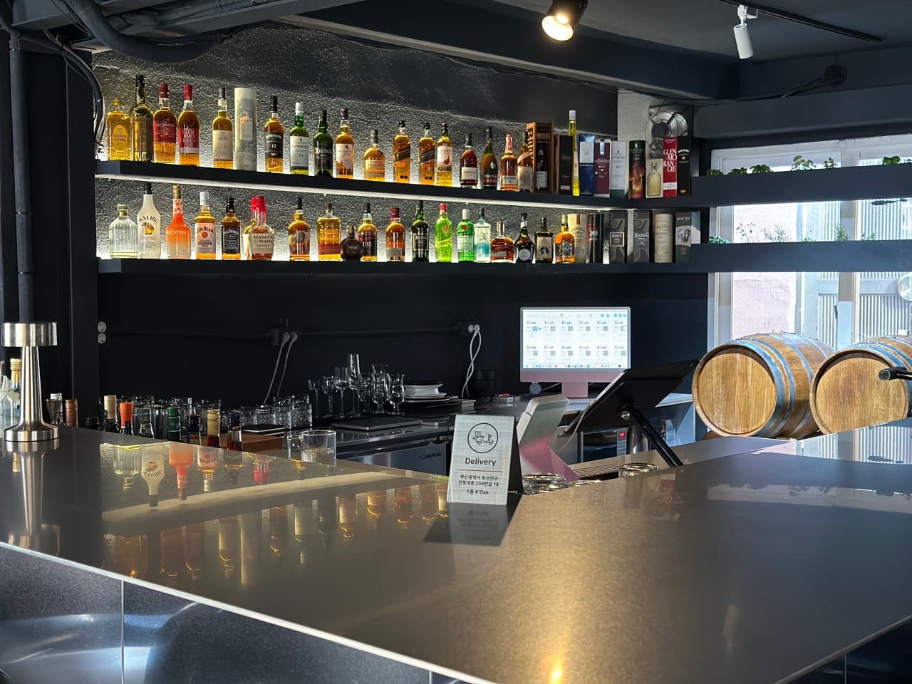 벽면 선반에 진열된 다양한 술병들과 조명을 반사하는 매끄러운 금속 재질의 바 카운터 좌석