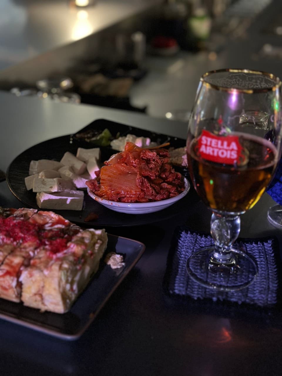어두운 조명의 바 테이블 위 맥주 한 잔과 김치, 두부, 계란말이가 차려진 1인용 안주 세트