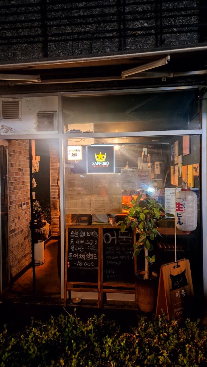 어두운 밤 통유리창 너머로 노란 조명이 비치고 입구에 메뉴 칠판과 일본식 제등이 배치된 바 테이블 중심의 소규모 매장 외부