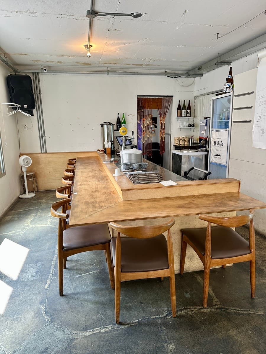 긴 L자형 나무 카운터 테이블과 일렬로 배치된 목재 의자가 있는 1인 중심의 바 좌석과 주방 내부 전경