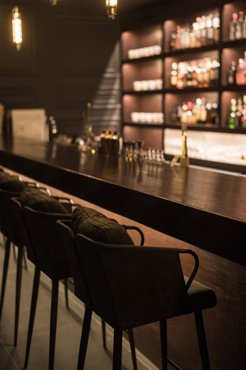 어두운 조명 아래 1인용 좌석이 일렬로 배치된 긴 목재 카운터와 다양한 술병이 진열된 선반
