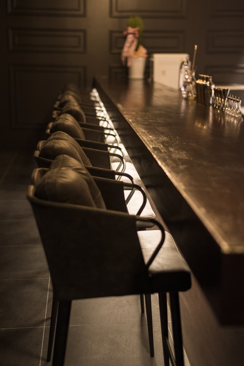 어두운 실내 조명 아래 길게 뻗은 나무 카운터 석과 일렬로 배치된 1인용 가죽 의자 및 바 도구들