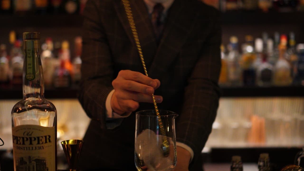 어두운 조명 아래 정장 차림의 바텐더가 바 카운터에서 긴 스푼으로 얼음을 젓는 모습과 위스키 병 및 지거