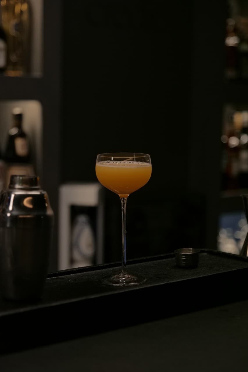 어두운 조명의 바 테이블 위 오렌지색 액체가 담긴 쿠페 글라스와 금속 소재의 칵테일 쉐이커