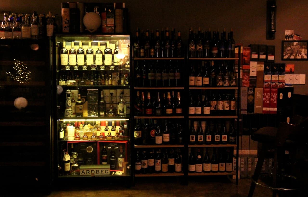 어두운 조명 아래 밝게 빛나는 양주 진열장과 벽면 선반을 가득 채운 술병들, 구석에 배치된 1인용 바 좌석