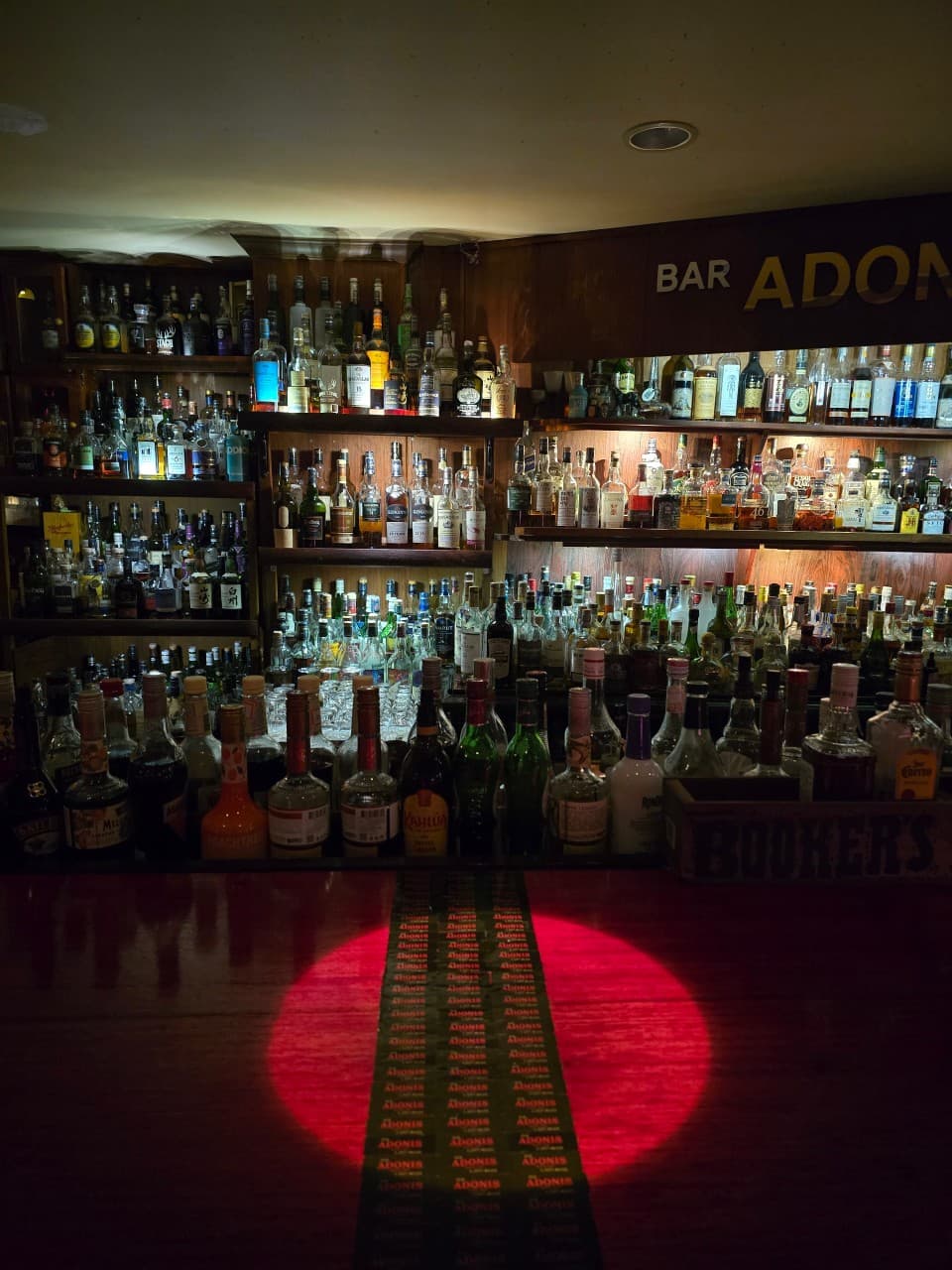 수많은 술병이 진열된 벽면 선반과 붉은색 조명이 내리쬐는 어두운 톤의 1인용 목재 바 테이블