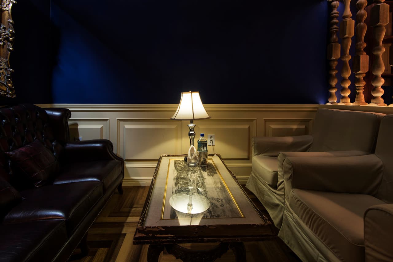 남색 벽면과 웨인스코팅 배경 아래 가죽 소파가 배치된 공간. 낮은 테이블 위에는 켜진 스탠드 조명과 생수병이 놓여 있다.