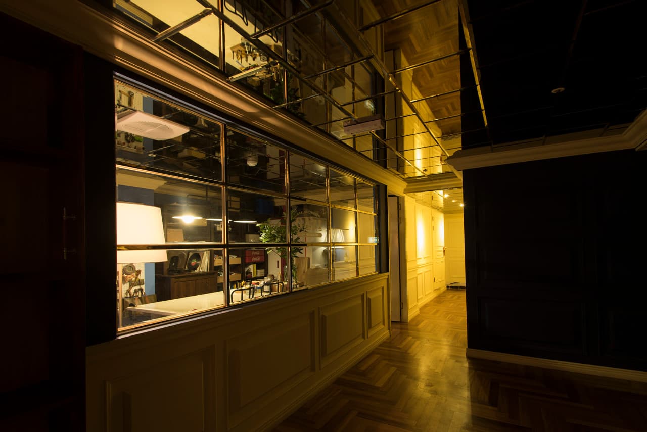 황색 조명이 비치는 헤링본 목재 바닥과 격자창 너머 바 테이블 공간. 거울로 마감된 천장 아래 어두운 벽면이 배치된 좁은 복도 통로