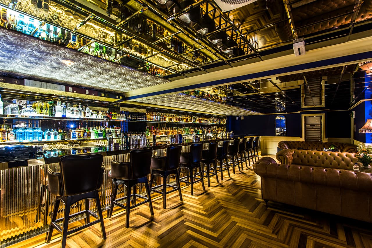 거울 천장 아래 수많은 술병이 진열된 긴 바 카운터와 일렬로 배치된 높은 가죽 의자들, 헤링본 패턴의 나무 바닥
