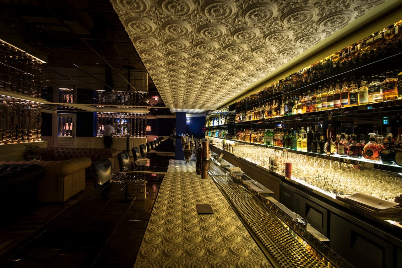 조명이 켜진 술병 선반 앞 양각 문양이 새겨진 긴 카운터와 일렬로 배치된 1인용 가죽 의자