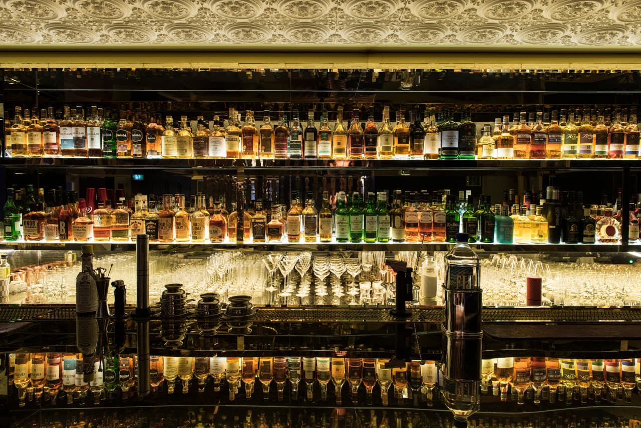 수많은 종류의 술병이 진열된 선반 아래 정렬된 유리잔과 반사되는 바 카운터의 긴 테이블