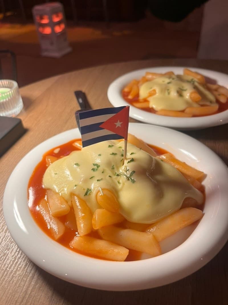 어두운 조명 아래 나무 테이블 위 치즈가 덮인 떡볶이와 작은 국기 장식이 꽂힌 흰색 접시