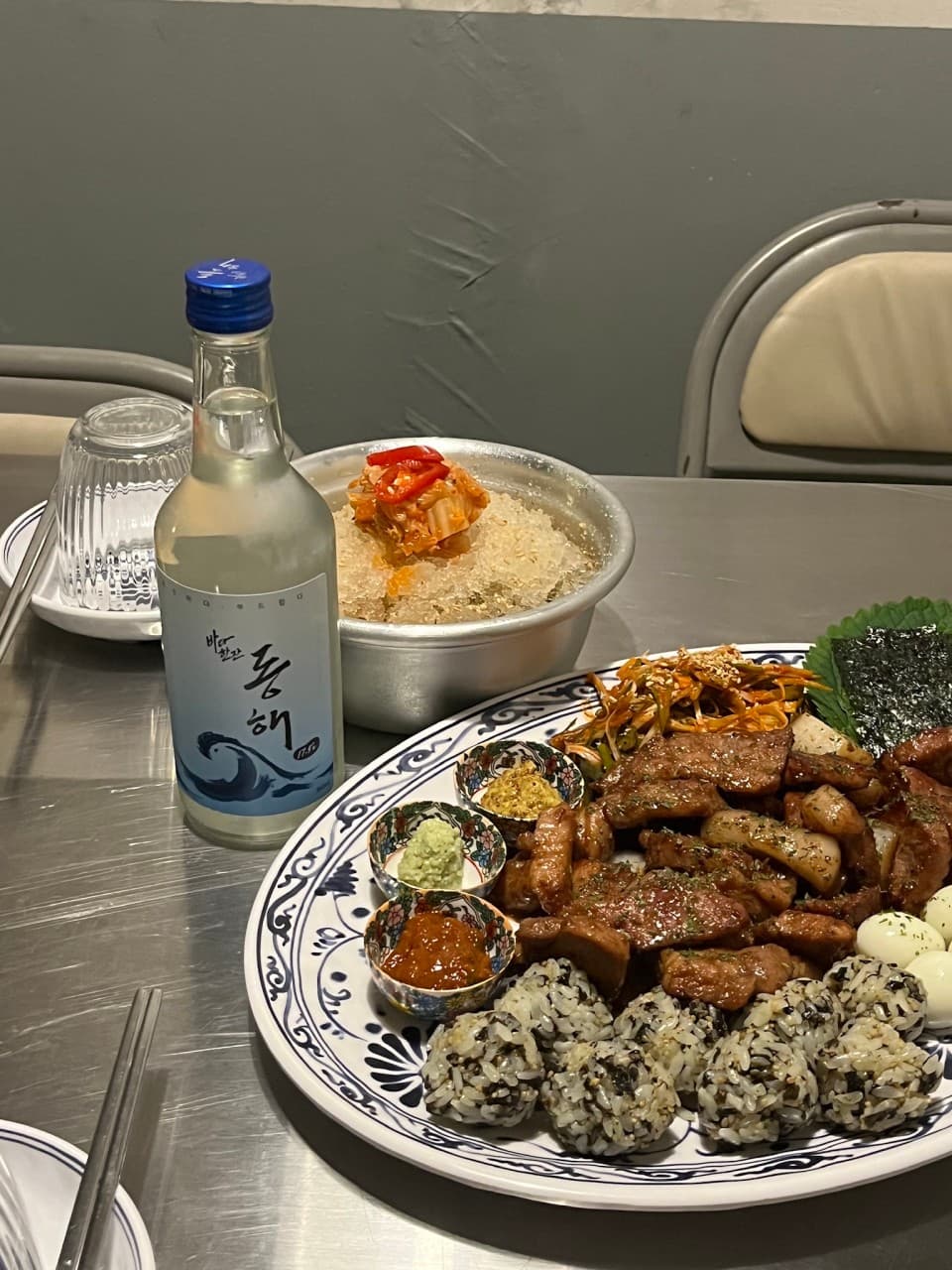 은색 금속 테이블 위 동해 소주병과 고기 구이, 주먹밥, 소스들이 담긴 접시와 살얼음 띄운 김치 요리