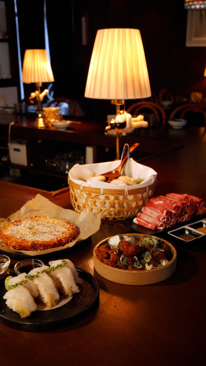 주황색 스탠드 조명이 비치는 어두운 목재 바 테이블 위 묵은지 초밥과 가라아게, 감자전과 얇게 썬 소고기가 차려진 전경