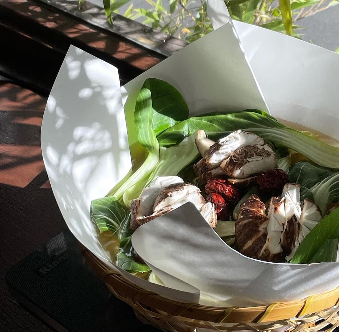 햇빛이 비치는 창가 좌석 위 대바구니에 담긴 1인용 종이 전골 냄비와 표고버섯, 청경채