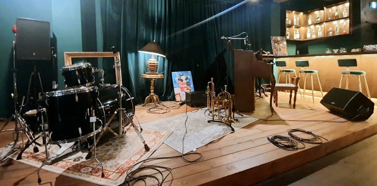 어두운 조명 아래 드럼과 트럼펫이 설치된 나무 무대와 1인용 바 좌석이 배치된 카운터 테이블 전경