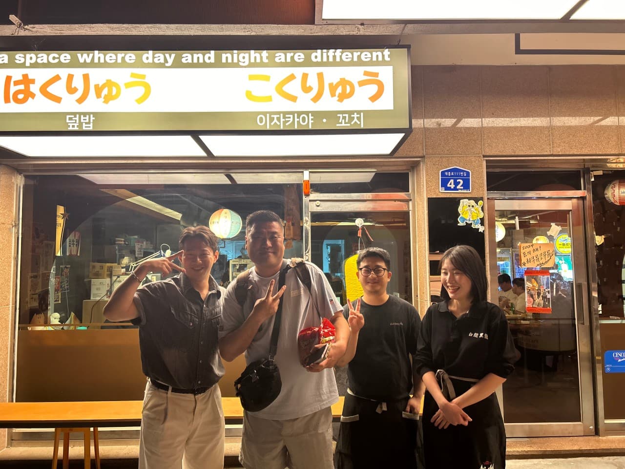 노란 조명 간판 아래 인물들이 선 식당 입구. 통유리창 너머로 1인석 바 테이블과 소품이 배치된 내부가 보이는 밤 거리의 이자카야 외관