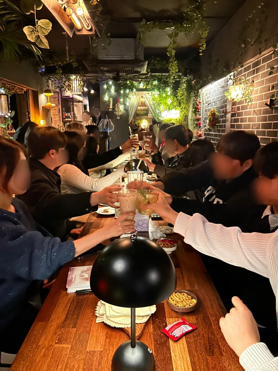 노란 조명 아래 긴 나무 카운터석에 나란히 앉아 하이볼 잔을 들어 올린 사람들과 벽면의 벽돌 장식