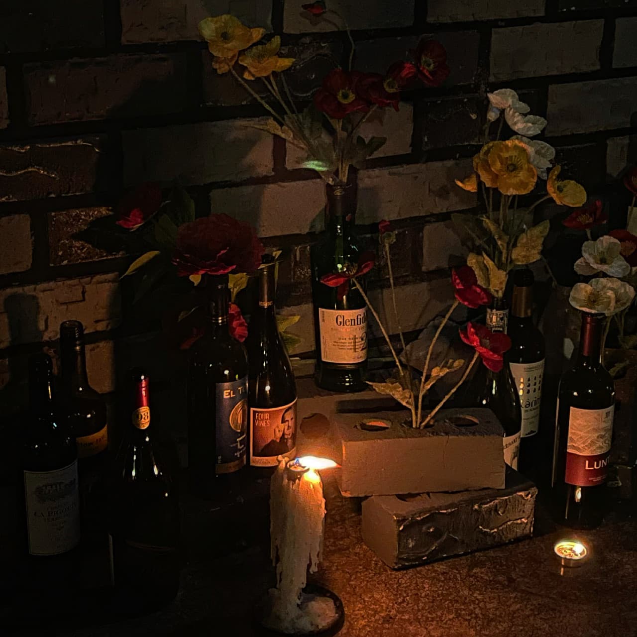 벽돌 벽 앞 촛농이 흘러내린 양초와 조화가 꽂힌 와인병들. 어두운 조명 아래 술병들이 나란히 놓인 바 테이블 전경