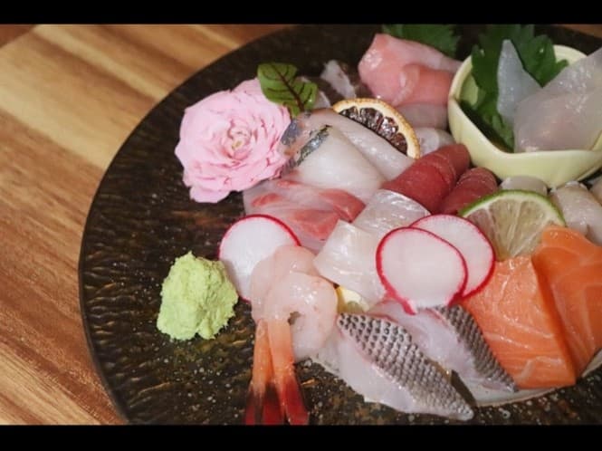 나무 질감 테이블 위 검은 원형 접시에 담긴 연어, 참치, 새우 사시미와 분홍색 꽃 장식 및 고추냉이