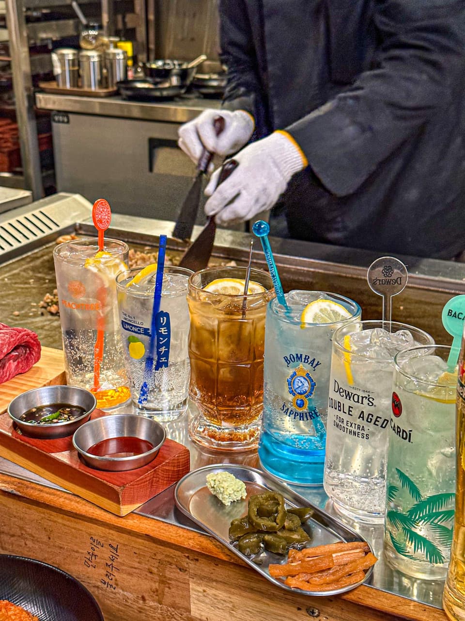 철판 앞 바 테이블에 정렬된 다양한 하이볼 잔들과 소스 접시 너머로 음식을 조리하는 요리사