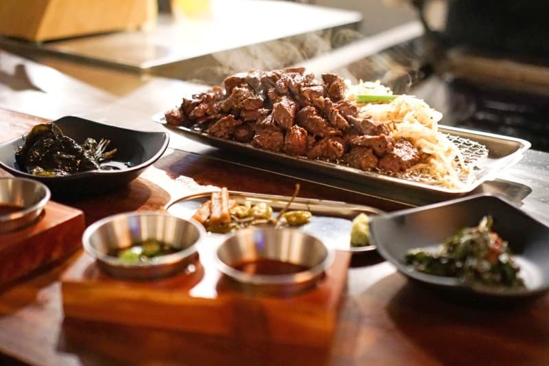 나무 소재의 바 테이블 위에 놓인 김이 나는 고기 요리와 숙주나물, 두 가지 소스가 담긴 1인용 상차림