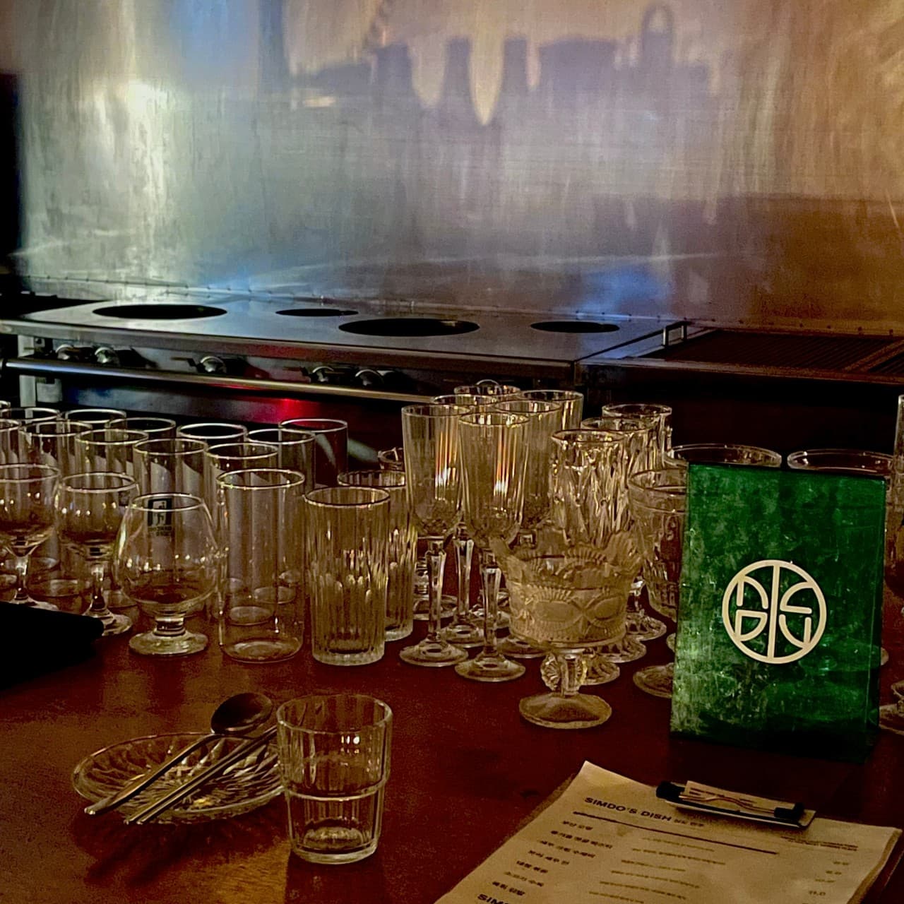 어두운 목재 바 테이블 위에 정렬된 다양한 유리잔과 1인용 수저가 놓인 접시, 메뉴판과 녹색 아크릴 소품 전경