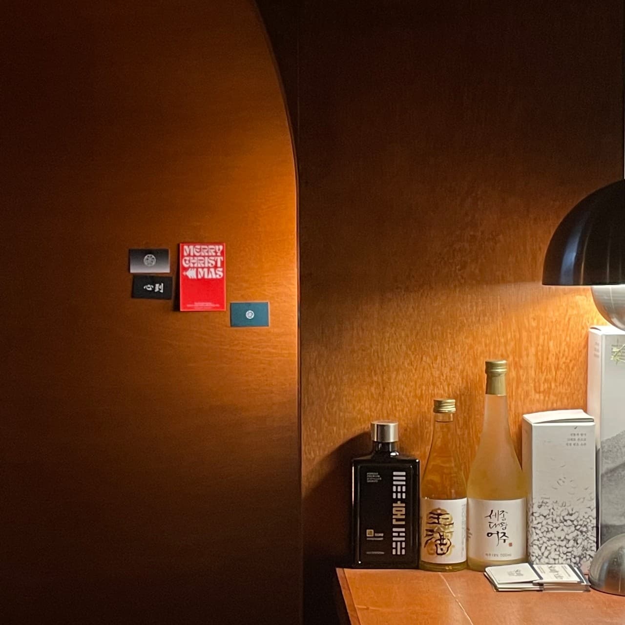 주황색 조명이 비추는 어두운 목재 벽면 앞 아치형 입구와 바 카운터 위 전통주 병들, 검은색 스탠드 조명