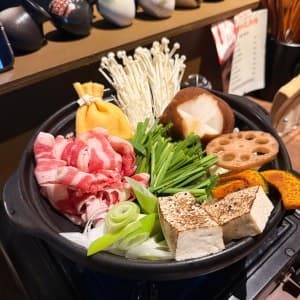 목재 카운터 위 휴대용 가스레인지에 놓인 소고기, 표고버섯, 유부주머니, 구운 두부가 담긴 검은색 전골 냄비