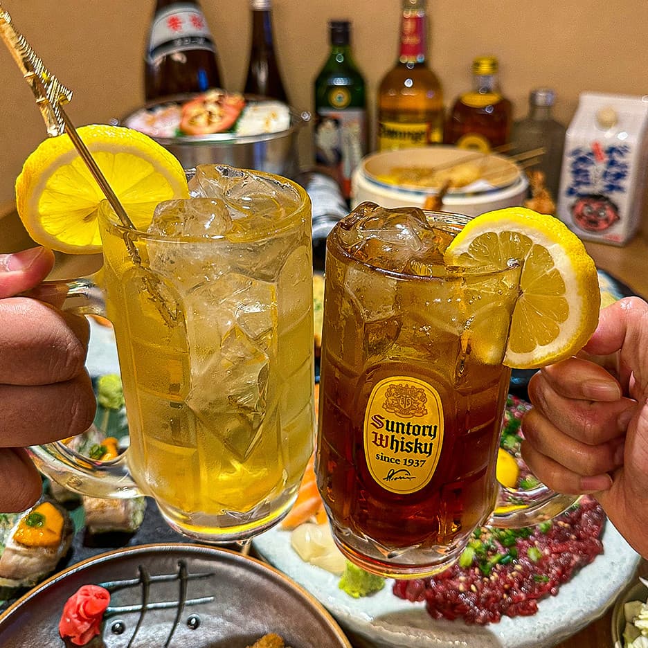 레몬 슬라이스를 곁들인 하이볼 두 잔과 테이블 위 육회, 연어 사시미 및 배경에 진열된 다양한 술병들