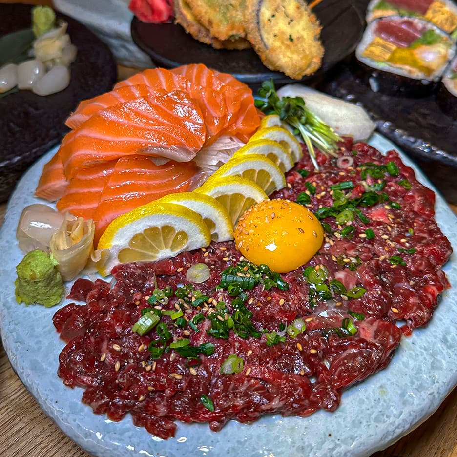 나무 테이블 위 연어 사시미와 노른자가 올라간 육회 한 접시, 레몬 슬라이스와 와사비 및 튀김