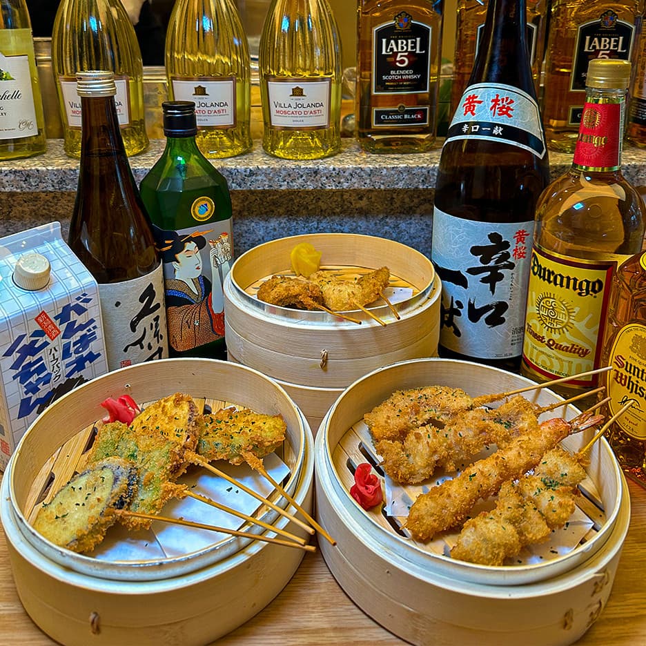 여러 종류의 튀김 꼬치가 담긴 세 개의 나무 찜기와 카운터 너머로 일렬로 늘어선 사케 및 위스키 병들